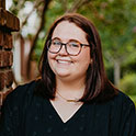 Kate is pictured from the shoulders up wearing a black shirt. She has brown hair, fair skin, glasses, and is smiling.