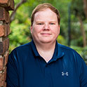 Andrew is pictured from the shoulders up. He is wearing a navy shirt, has blonde hair and fair skin and is smiling.