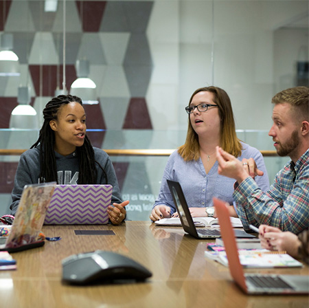 Three colleagues seated around a conference table having a discussion, each with an open laptop. One woman is speaking while the others listen.