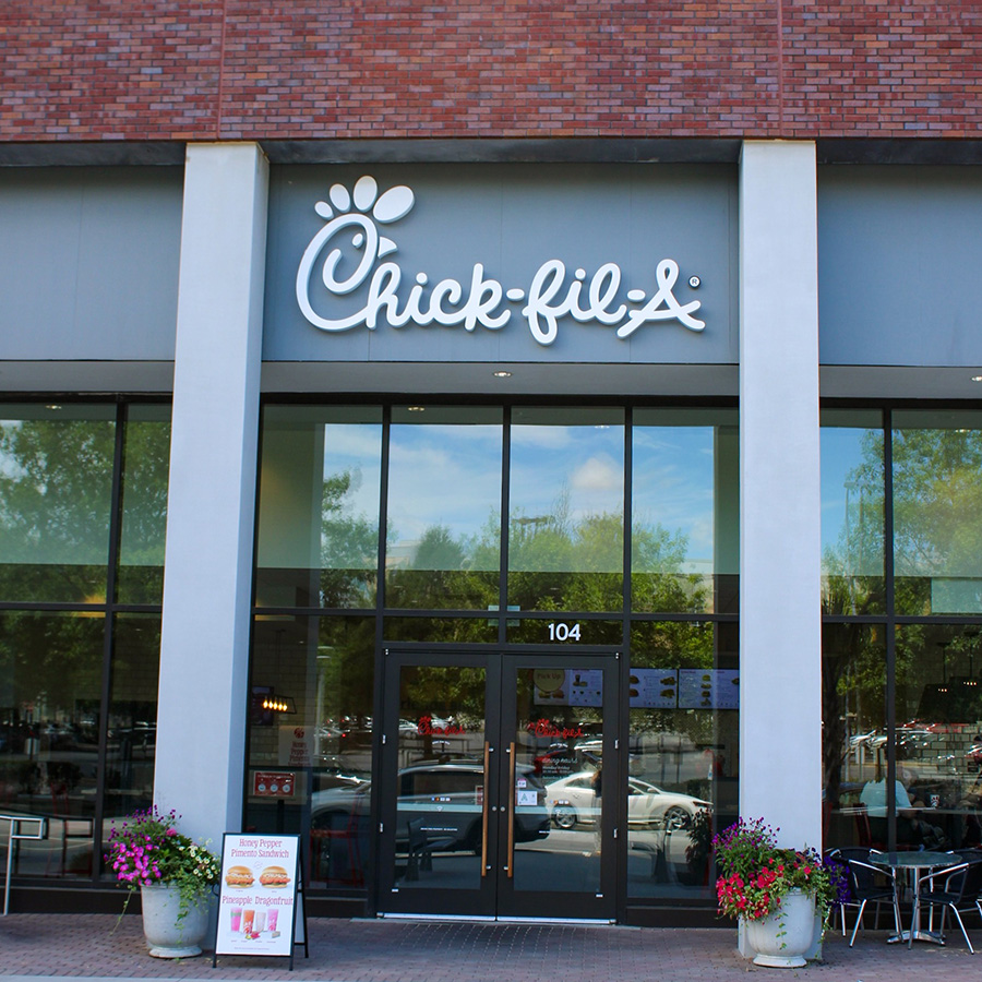 Exterior entrance of a Chick-fil-A restaurant with large glass windows, outdoor seating, and flower planters.