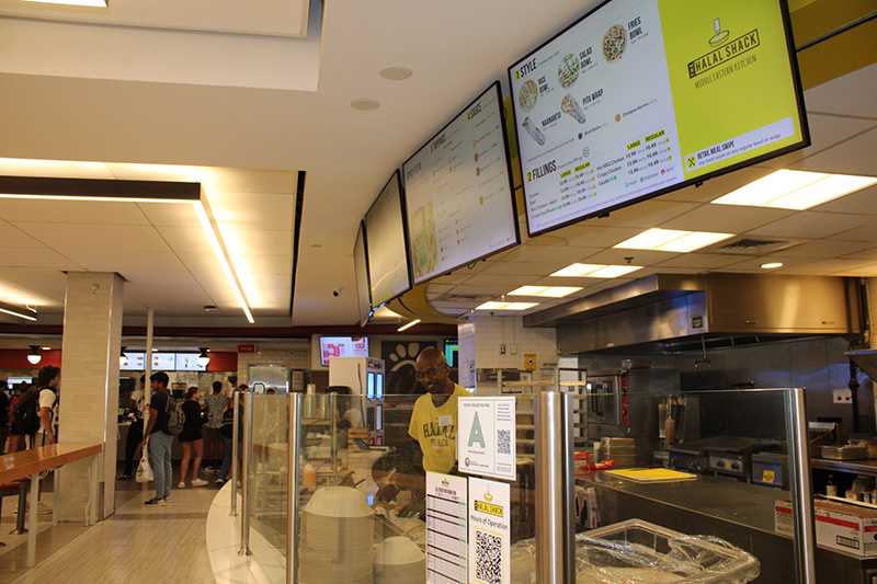 Halal Shack food counter in a campus dining hall with digital menu boards, and an employee behind the counter.