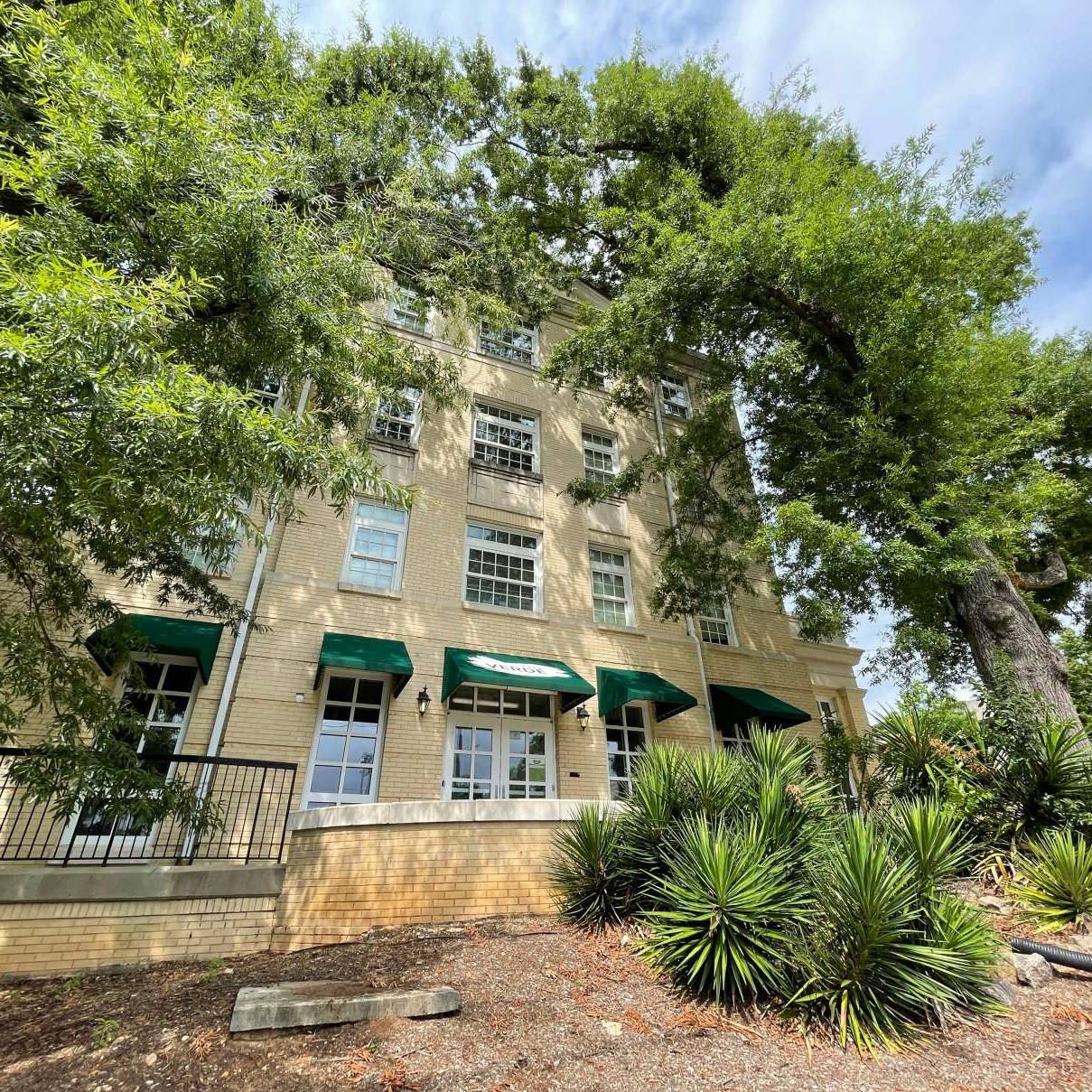 Multi-story brick building with green awnings, surrounded by large shade trees and landscaped greenery.