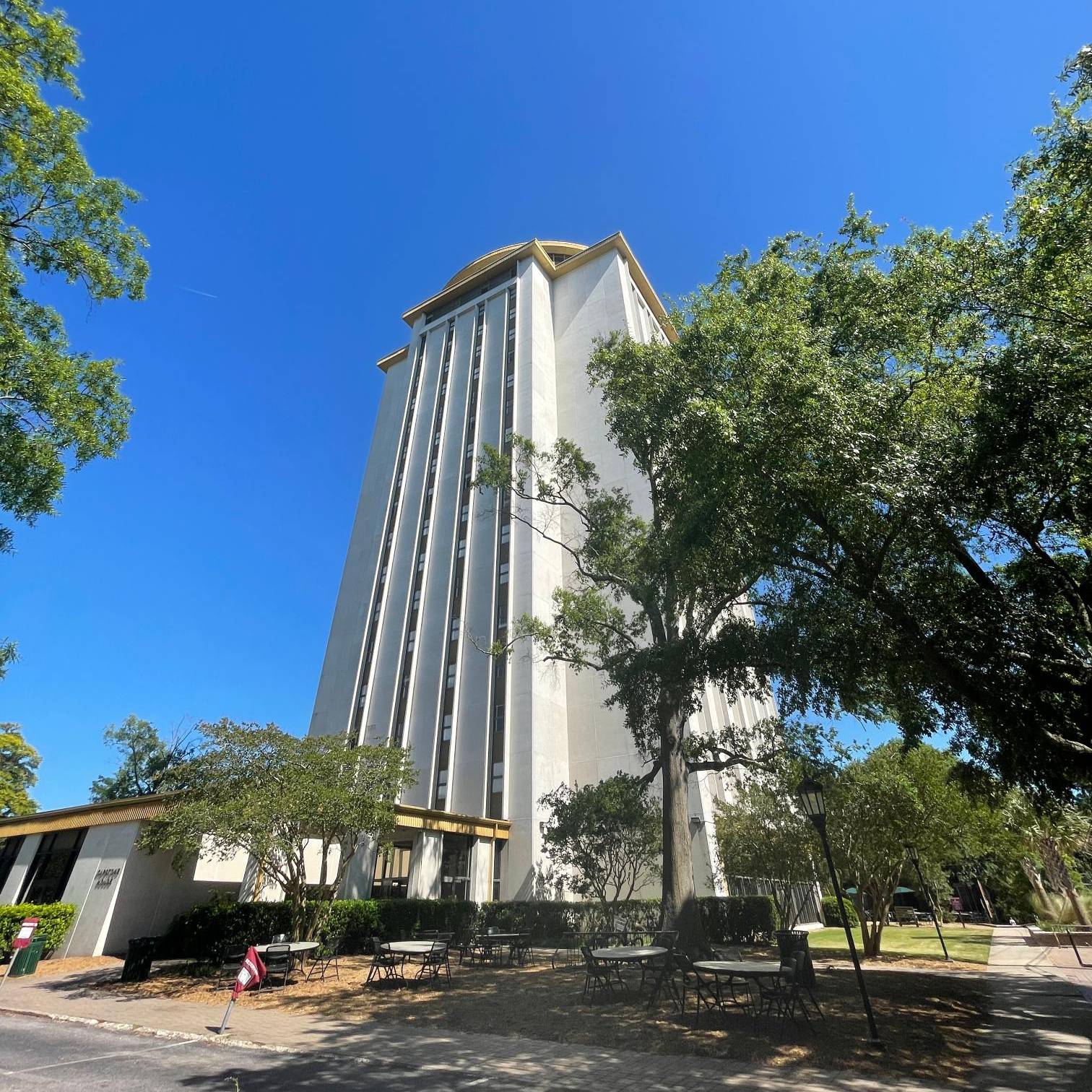 Tall campus residence tower surrounded by mature trees, with outdoor tables and chairs beneath a bright blue sky.