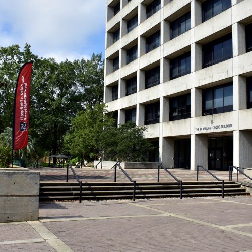 Exterior of the University of South Carolina’s T. W. Gambrell Close Building, with a wide staircase, trees, and a red Hospitality, Retailing and Sport Management banner.