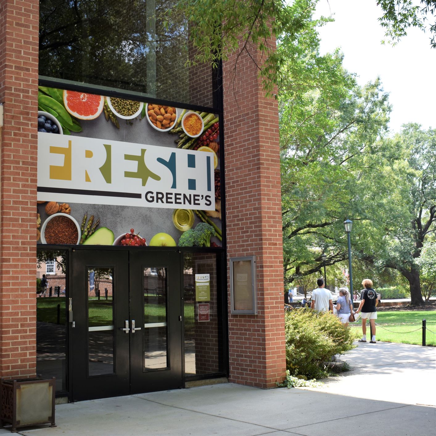 Entrance to Fresh Greene’s in a brick campus building, with a large food-themed sign and a tree-lined walkway nearby.