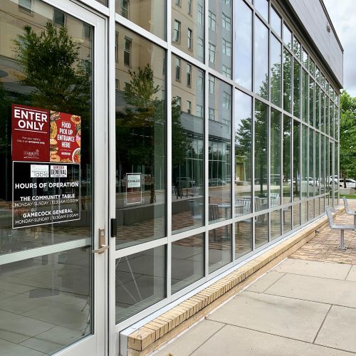 Glass entrance to a campus dining location with posted entry, food pickup, and hours-of-operation signs.