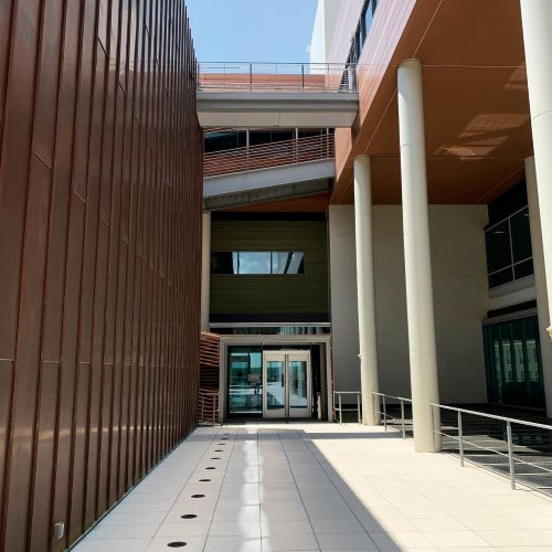 Modern building courtyard with tall columns, metal-paneled walls, a tiled walkway, and glass entrance doors.