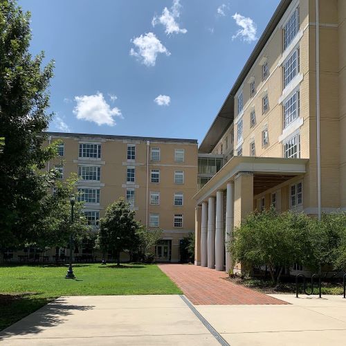 Multi-story campus residence buildings surrounding a green lawn and brick walkway under a bright blue sky.