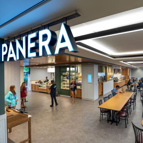 Panera dining location with a large illuminated sign, ordering counter, communal tables and customers inside.