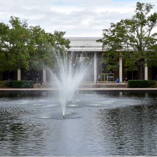 Large fountain spraying water in a reflecting pool surrounded by trees, with a campus building in the background.