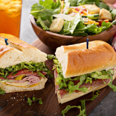 Two deli sandwiches with lettuce, tomato, onion, and sliced meats served on a wooden board with a fresh green salad.