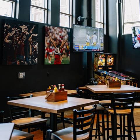 Casual sports-themed dining area with wooden tables and chairs, wall-mounted TVs, athlete photos, and a pinball machine.