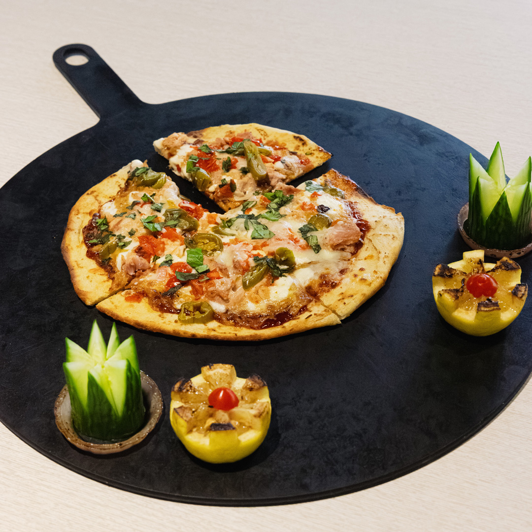 Flatbread pizza topped with chicken, jalapeños, melted cheese, tomatoes, and fresh herbs, served on a black platter with cucumber and grilled lemon garnishes.