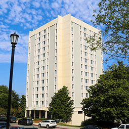 Exterior view of a tall, modern residence hall with a light gray and white facade rising above green trees under a clear blue sky. The multi-story building features vertical rows of windows and clean architectural lines, photographed from ground level on a bright, sunny day.