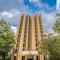 Exterior view of a tall campus residence hall with a light tan brick façade and narrow vertical rows of windows extending the height of the building. Mature trees frame the residence hall on both sides, and a decorative campus light is visible in the foreground. The building is photographed from ground level beneath a partly cloudy blue sky, creating a welcoming campus setting.