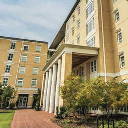 Exterior view of a modern campus residence hall with a light tan brick façade, large windows, and a covered entrance supported by tall white columns. A brick walkway leads through landscaped grounds with trees and shrubs, while bicycle racks and parked bicycles sit near the entrance. The building is photographed on a bright day beneath a blue sky with scattered clouds.