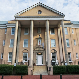 Exterior view of a historic campus residence hall with a light tan façade, a grand entrance supported by tall classical columns, and a triangular pediment above the front doors. A brick walkway leads to the building, which is framed by mature trees and landscaped hedges beneath a partly cloudy sky, creating a stately and welcoming campus setting.