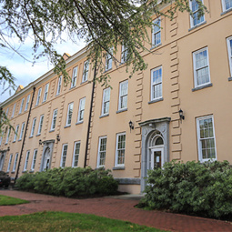 Exterior view of Woodrow College Residence Hall, a historic multi-story campus residence building with a light tan façade, tall windows, and classical architectural details. Trees, shrubs, and a brick walkway frame the building on a bright day, creating a welcoming campus setting.
