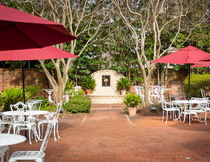 Brick patio with white tables and chairs and red umbrellas and a fountain in the back with a plaque with a palmetto tree on it. 