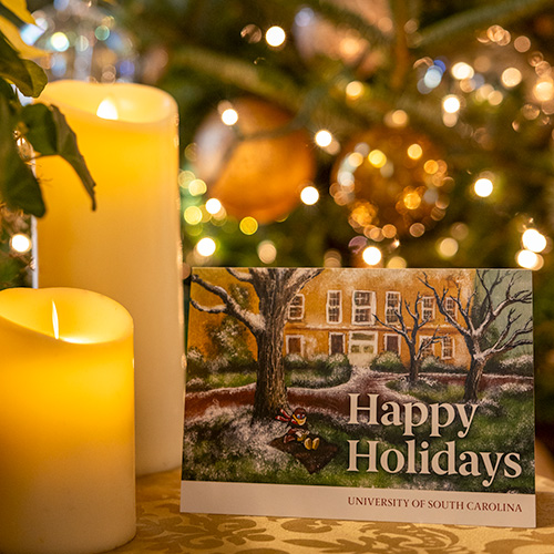 A tabletop with candles and greenery highlighting USC’s 2025 holiday card featuring an illustration of Cocky sitting on the Horseshoe