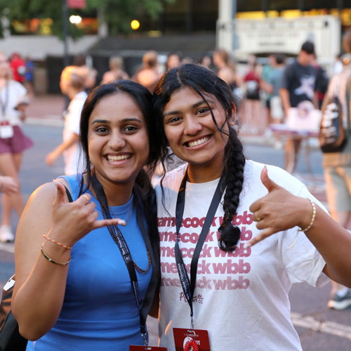 Two college students smile at the camera outdoors, both making spurs up hand gestures.