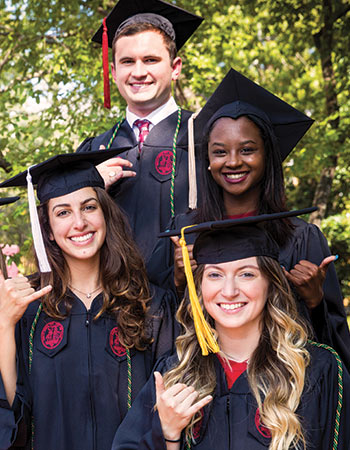 Four students in graduation cap and gowns smiling at the camera holding up the spurs up gesture.