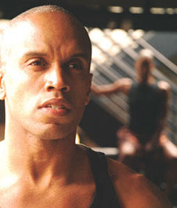 a man standing looking onward with a shaved head and a blurred view of dancers in the background