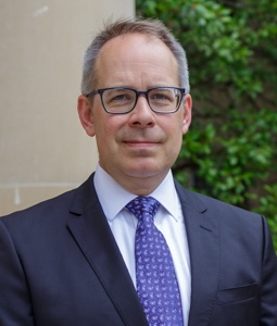 Carl Dahlman has short gray hair and black framed glasses, and wears a navy blazer, white shirt, and patterned blue tie.