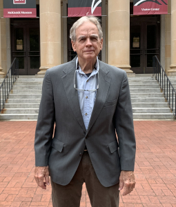 Don Jordan wears a gray blazer and blue button down shirt with his glasses around his neck. He stands with his arms to the side in front of McKissick Museum's steps and columns.