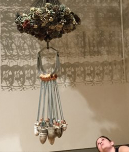 A woman looks up at a hanging art installation. The piece features a cluster of plants or natural materials at the top, with straps suspending several acorn-shaped objects below. The installation casts intricate shadows on the wall behind it.