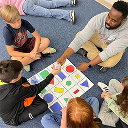 Several children and a man sit around a mat with colored shapes. The man passes a child a block.