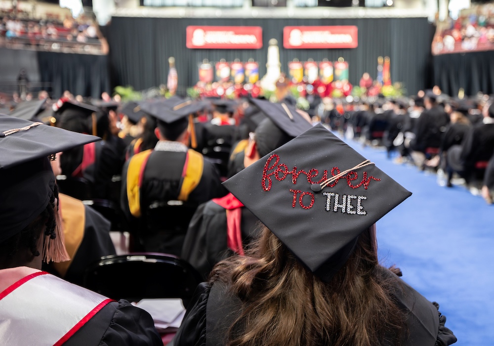 woman sitting in crowd wearing black graduation cap with glittery lettering that reads 'forever to thee'