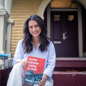 Woman sits on a porch and holds a book titled Another Sojourner Looking for Truth by Millicent E. Brown.