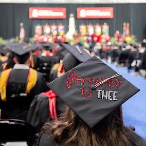 woman sitting in crowd wearing black graduation cap with glittery lettering that reads 'forever to thee'
