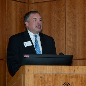 man wearing black suit and blue tie speaking behind brown lectern