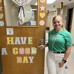Sophia Comose stands next to a decorated classroom door that reads, 'Have A Good Day.'