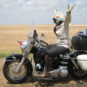 Kay Andrea rides a vintage motorcycle along Route 66, her hands raised in the air